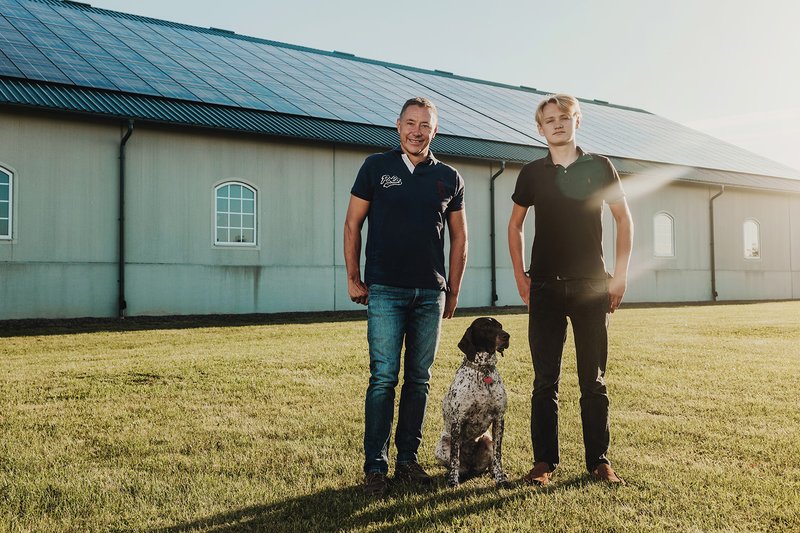 Ein Vater und ein Sohn mit einem Hund vor ihrer Scheune, welche mit PV-Anlagen ausgestattet ist (Solarpaket I: Wie profitieren Bürgerinnen und Bürger davon?).